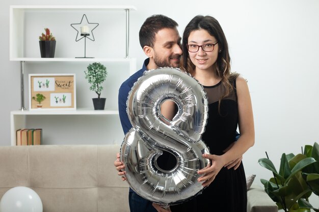 Hombre guapo complacido mirando a una mujer joven y bonita con gafas ópticas sosteniendo un globo en forma de ocho de pie en la sala de estar en el día internacional de la mujer de marzo