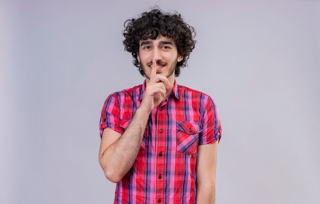 Un hombre guapo complacido con el pelo rizado en camisa a cuadros manteniendo el dedo índice en la boca