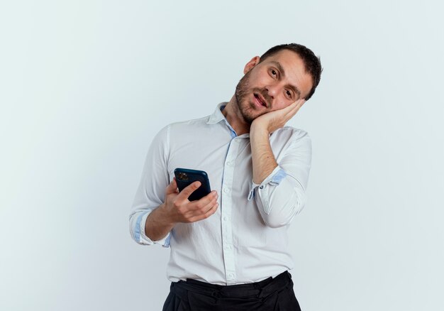 Hombre guapo complacido pone la mano en la cara sosteniendo el teléfono aislado en la pared blanca