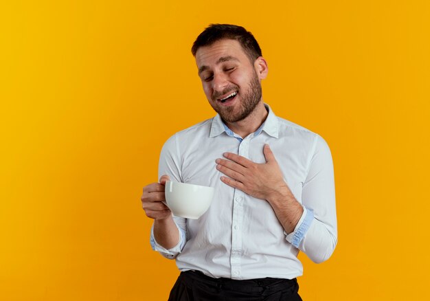 Hombre guapo complacido pone la mano en el pecho sosteniendo la taza aislada en la pared naranja