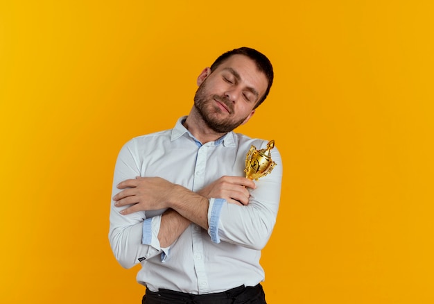 Hombre guapo complacido sostiene y abraza la copa ganadora aislada en la pared naranja