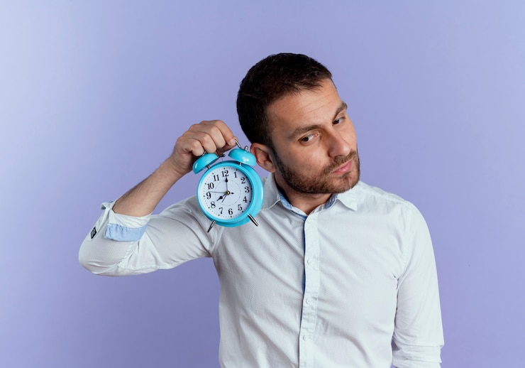 Foto gratuito hombre guapo complacido sostiene el reloj de alarma cerca de la oreja tratando de escuchar aislado en la pared púrpura