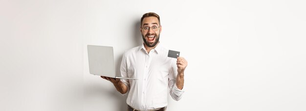 Hombre guapo de compras en línea que muestra la tarjeta de crédito y usa una computadora portátil para ordenar en internet de pie