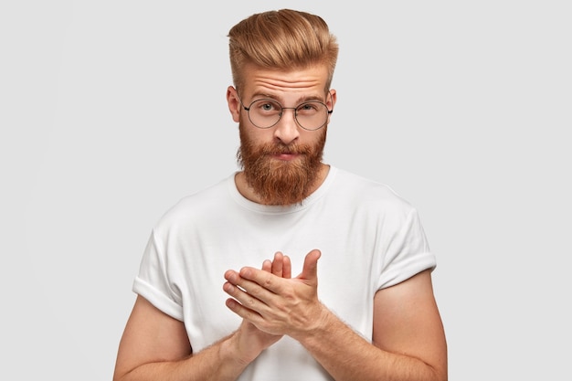 Hombre guapo confiado aplaude cuando saluda a alguien, tiene una espesa barba pelirroja y un corte de pelo moderno, vestido informalmente, aplaude con ambas manos, aislado sobre una pared blanca. Concepto de personas y felicitaciones