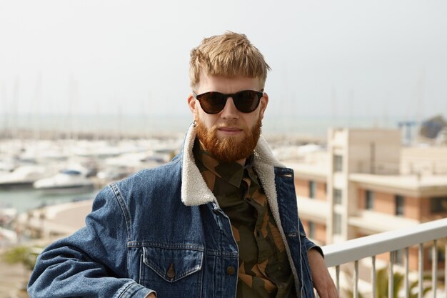 Hombre guapo confiado con barba borrosa de pie en el punto de vista con los brazos sobre los rieles de la cerca blanca