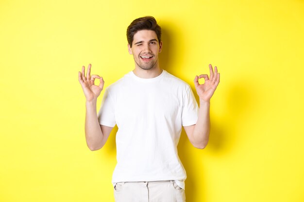 Hombre guapo confiado guiñando un ojo, mostrando signos de aprobación en señal de aprobación, como algo bueno, de pie sobre fondo amarillo