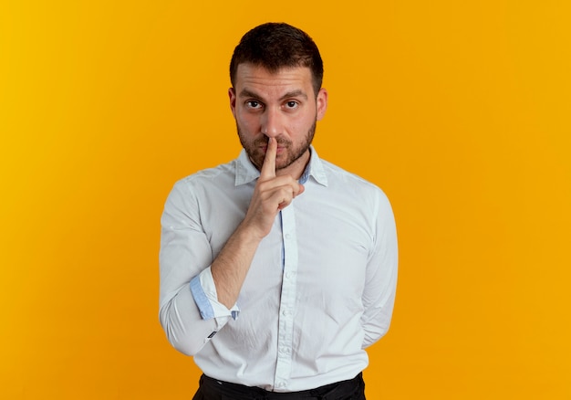 Hombre guapo confiado pone el dedo en la boca gesticulando silencio tranquilo aislado en la pared naranja