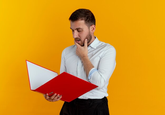 Hombre guapo confiado pone la mano en la barbilla mirando la carpeta de archivos aislada en la pared naranja