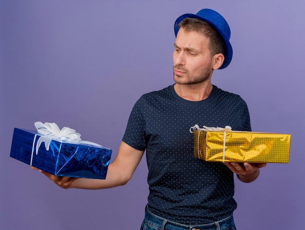 Hombre guapo confundido con sombrero azul sostiene y mira cajas de regalo aisladas en la pared púrpura