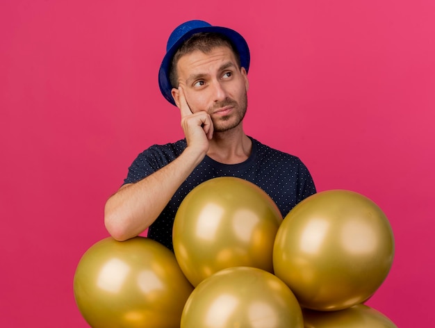 Hombre guapo confundido con sombrero de fiesta azul sostiene globos de helio mirando al lado aislado en la pared rosa con espacio de copia