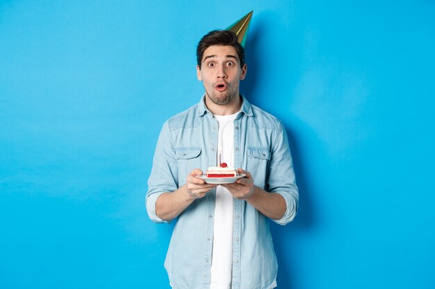 Hombre guapo en cono de fiesta sosteniendo pastel de cumpleaños, mirando sorprendido, de pie sobre fondo azul.