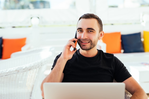 Hombre guapo en cubierta con teléfono móvil y computadora portátil en zona de salón de resort de verano