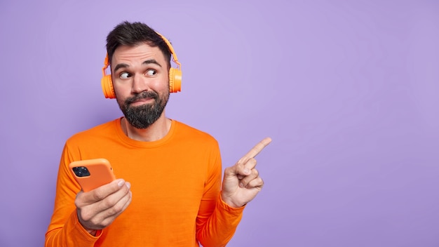 Hombre guapo curioso con barba demuestra puntos interesantes en el espacio en blanco en la pared púrpura usa el teléfono móvil para chatear en línea y escucha música usa auriculares vestidos de manera informal