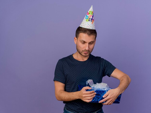 Hombre guapo decepcionado con gorro de cumpleaños tiene caja de regalo aislada en la pared púrpura con espacio de copia