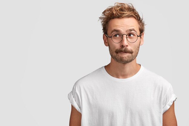 Hombre guapo desconcertado con bigote, muerde el labio inferior y mira con curiosidad a un lado, piensa en algo, vestido con una camiseta blanca informal, se para contra la pared en blanco