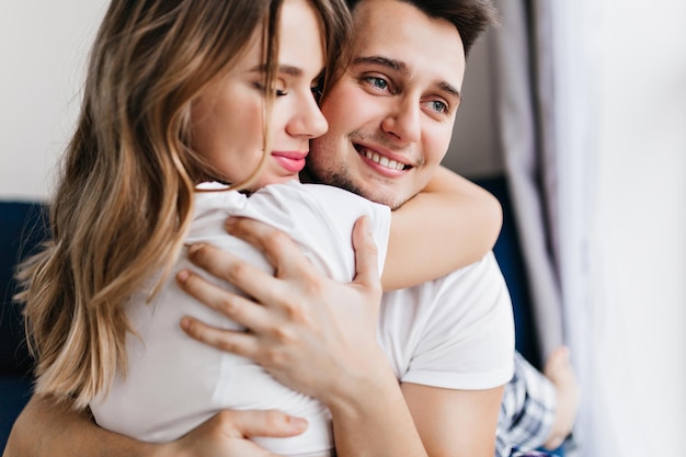 Hombre guapo dichoso abraza a su novia. Retrato de interior de matrimonio europeo disfrutando de la mañana del fin de semana.