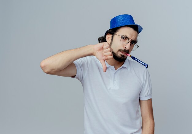 Hombre guapo disgustado con gafas y sombrero azul que sopla el silbato con el pulgar hacia abajo aislado en blanco