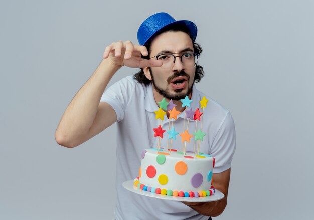 Hombre guapo disgustado con gafas y sombrero azul sosteniendo pastel aislado en blanco