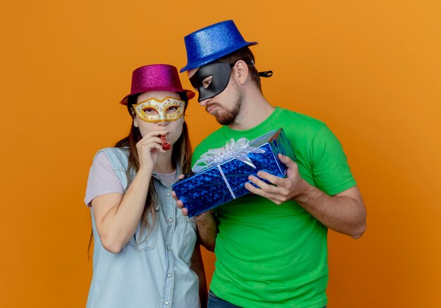 Hombre guapo disgustado con sombrero azul con máscara de ojos de mascarada con caja de regalo mirando a una joven alegre con sombrero rosa y máscara de ojos de mascarada que sopla silbato mirando al frente