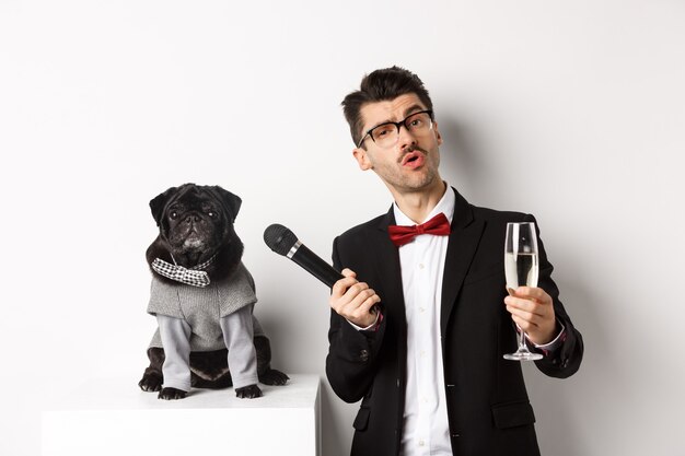 Hombre guapo elegante en vasos, levantando una copa de champán y dando el micrófono al pug lindo en traje de fiesta, celebrando y divirtiéndose, fondo blanco.