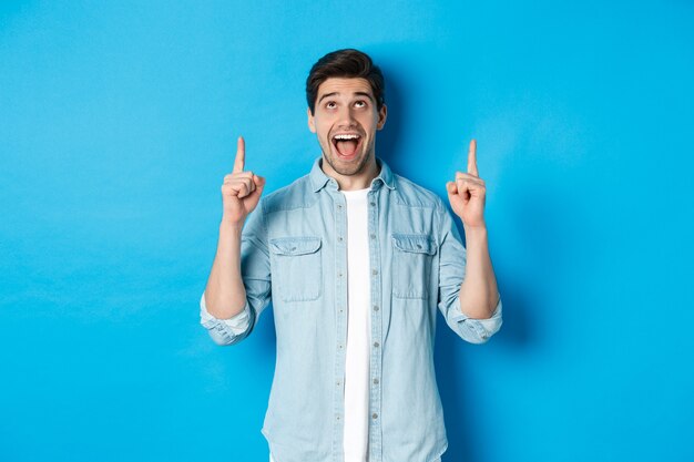 Hombre guapo emocionado apuntando y mirando hacia arriba, mostrando publicidad, de pie sobre fondo azul