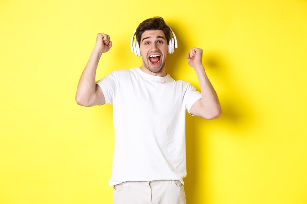 Hombre guapo emocionado bailando y cantando, escuchando música en auriculares, de pie sobre fondo amarillo