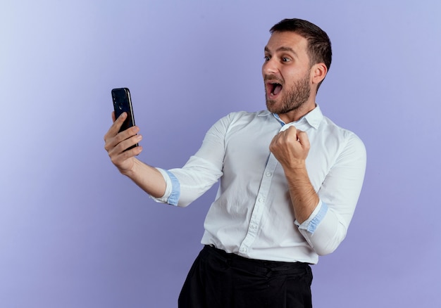 Hombre guapo emocionado mira el teléfono y mantiene el puño aislado en la pared púrpura