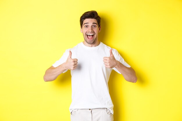 Hombre guapo emocionado mostrando los pulgares para arriba aprobar y decir que sí de pie sobre fondo amarillo