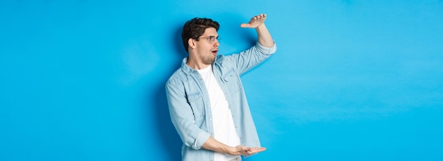 Foto gratuita un hombre guapo emocionado que muestra un objeto de gran tamaño y se ve asombrado de pie sobre un fondo azul