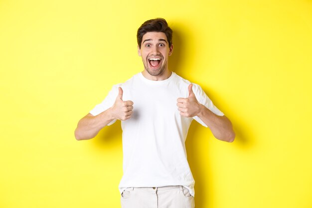 Hombre guapo emocionado que muestra los pulgares para arriba, aprueba y dice que sí, de pie sobre fondo amarillo.