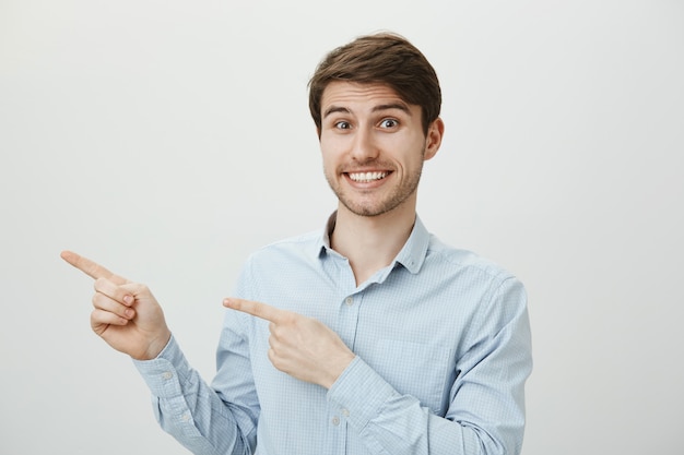 Hombre guapo emocionado sonriendo ampliamente, señalando con el dedo a la izquierda en el banner promocional