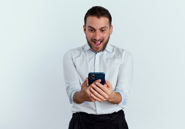 Hombre guapo emocionado sostiene y mira el teléfono aislado en la pared blanca