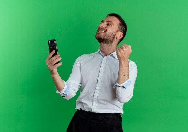 Hombre guapo emocionado sostiene el teléfono y mantiene el puño mirando hacia arriba aislado en la pared verde