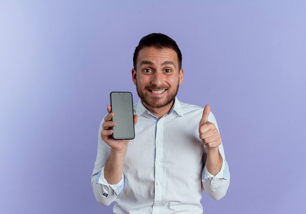 Hombre guapo emocionado sostiene teléfono Thumbs up aislado en la pared púrpura