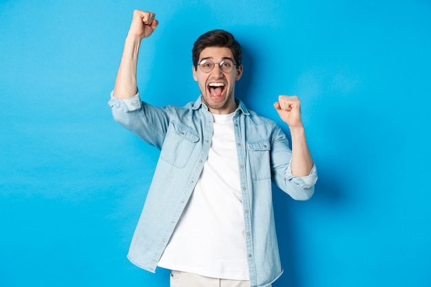 Foto gratuita hombre guapo emocionado triunfando, levantando las manos y gritando de alegría, celebrando la victoria, de pie contra el fondo azul