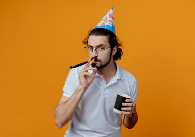 Hombre guapo enojado en gorro de cumpleaños soplando un silbato y sosteniendo una taza de café aislado sobre fondo naranja