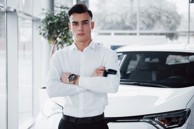 Un hombre guapo es un comprador parado junto a un auto nuevo en el centro de distribución y mirando a la cámara.