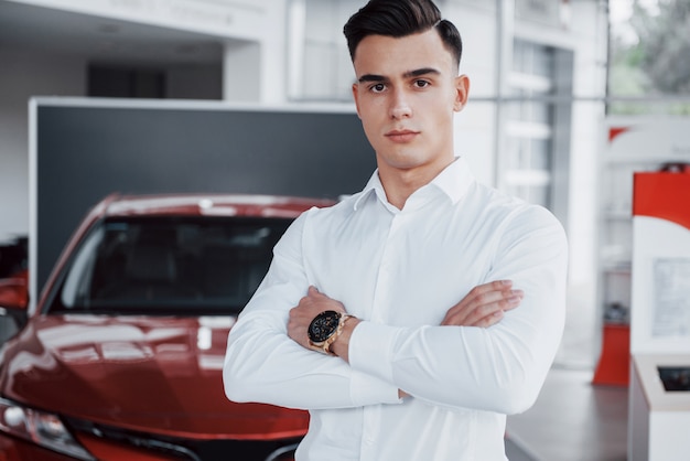 Un hombre guapo es un comprador parado junto a un auto nuevo en el centro de distribución y mirando a la cámara.