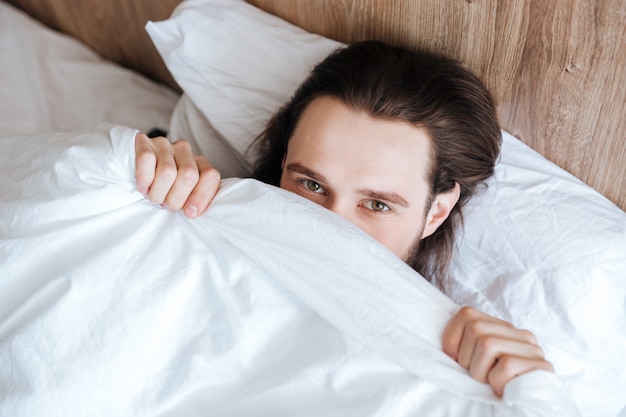 Hombre guapo escondiendo su rostro debajo de la colcha blanca en la cama