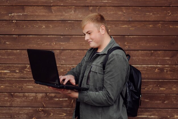 Hombre guapo está usando una computadora portátil y un teléfono al aire libre en la pared de madera