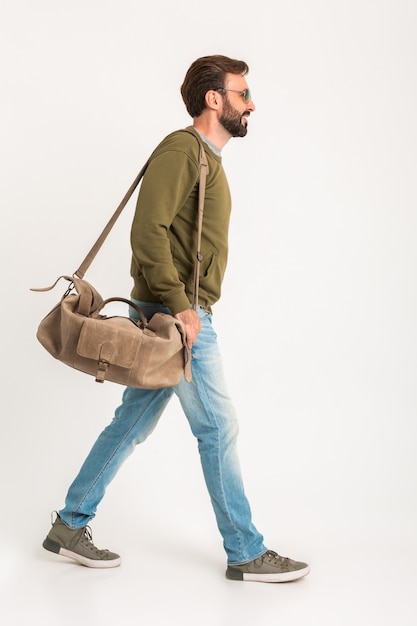 Hombre guapo con estilo barbudo caminando aislado vestido con sudadera con bolsa de viaje, jeans y gafas de sol