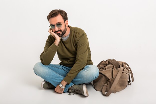 Hombre guapo con estilo barbudo sentado en el piso aislado vestido con sudadera con bolsa de viaje, jeans y gafas de sol, esperando triste y cansado