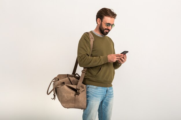 Hombre guapo con estilo barbudo en sudadera con bolsa de viaje, jeans y gafas de sol aislados sosteniendo el teléfono