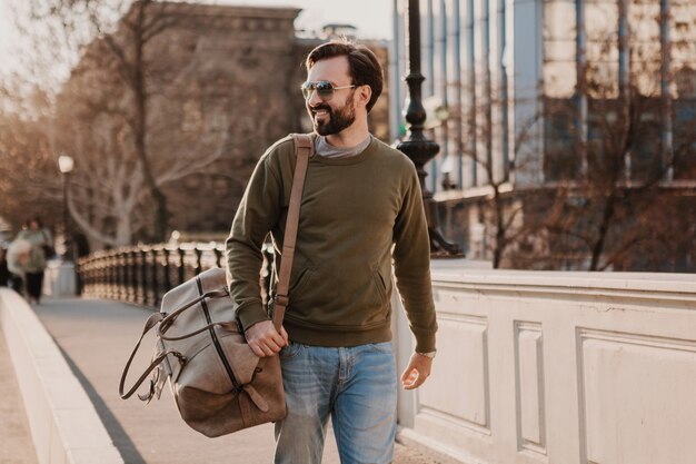 Hombre guapo con estilo hipster caminando en la calle de la ciudad con bolso de cuero con sudadera y gafas de sol, tendencia de estilo urbano, día soleado, viajero feliz sonriente