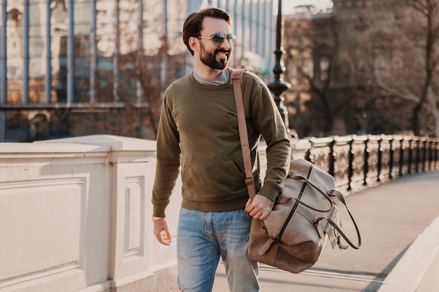 Hombre guapo con estilo hipster caminando en la calle de la ciudad con bolso de cuero con sudadera y gafas de sol, tendencia de estilo urbano, día soleado, viajero feliz sonriente