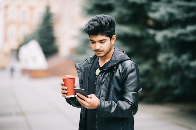 Hombre guapo estudiante con anteojos hablando con amigos por teléfono móvil disfrutando de café urbano mientras camina en la calle