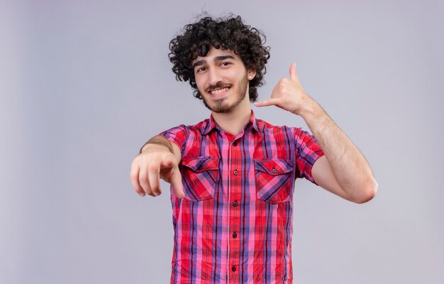 Un hombre guapo feliz con el pelo rizado en camisa a cuadros sosteniendo la mano cerca de la oreja, señal de teléfono de gesto con los dedos