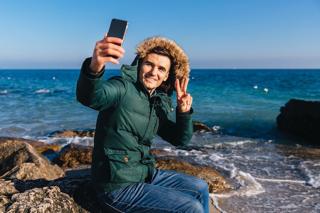 Hombre guapo feliz toma un selfie en smartphone, mostrando gesto de paz