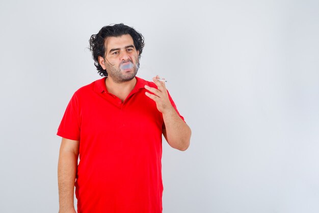 Hombre guapo fumando cigarrillo en camiseta roja y mirando serio. vista frontal.