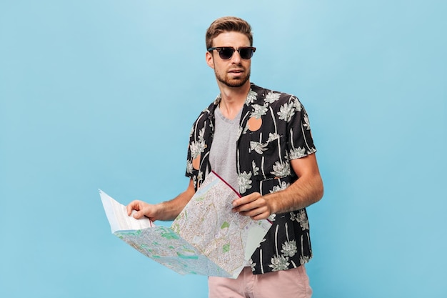 Hombre guapo con gafas de sol negras en camisa fresca de verano y camisa ligera mirando hacia otro lado y sosteniendo un mapa en un fondo aislado